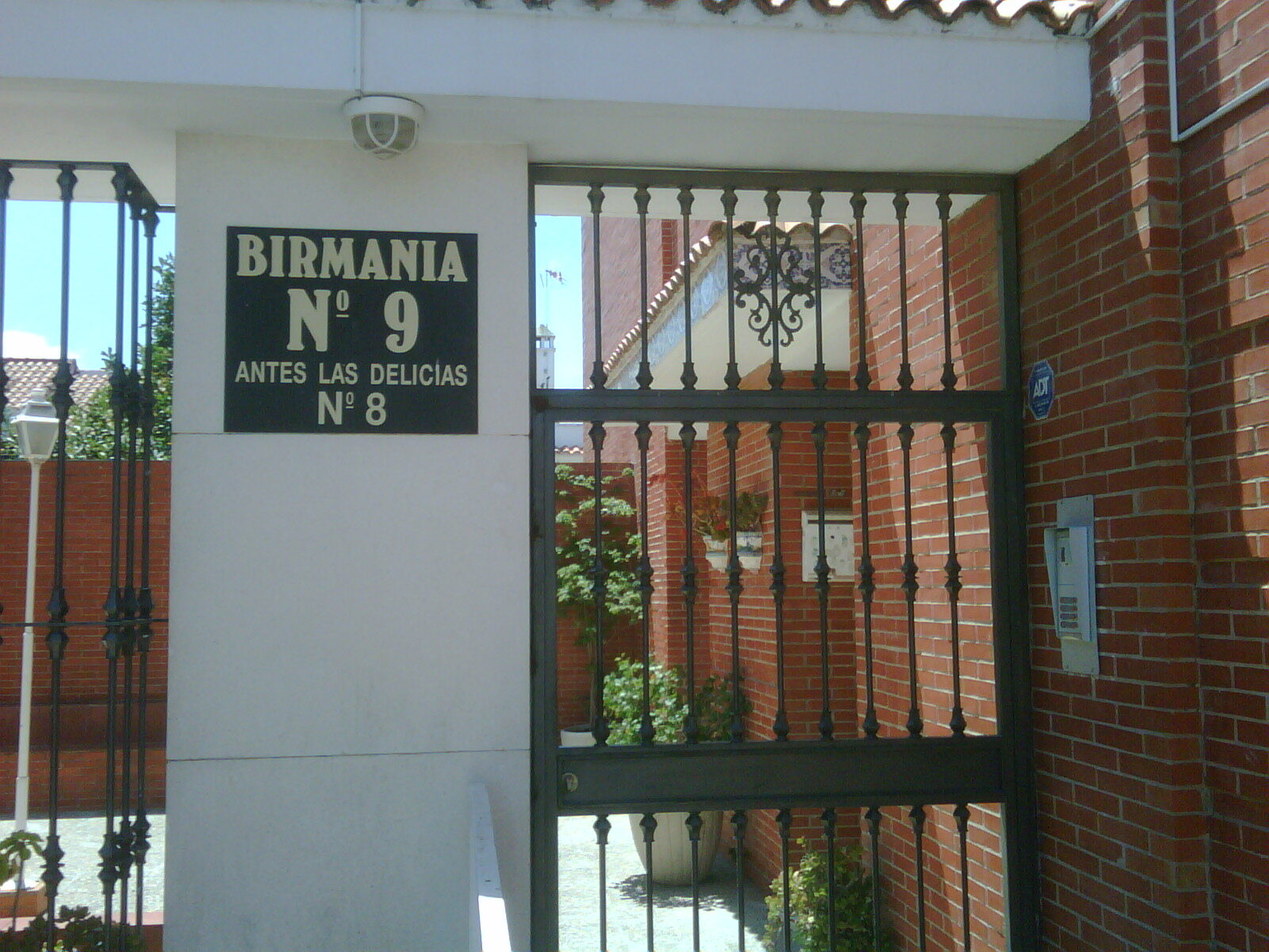 Puerta de hierro negro con diseño ornamental, cerrada y enmarcada por un entorno ajardinado.